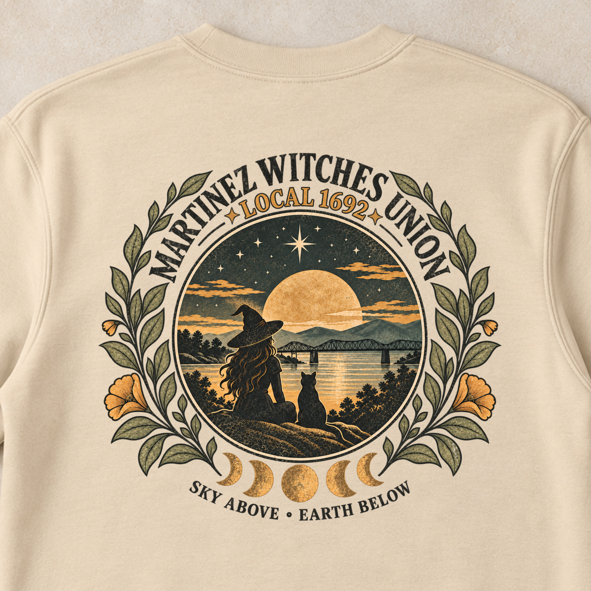 A cream-colored crewneck sweatshirt with a large graphic print on the back, laid flat on a light beige textured surface with small decorative pumpkins and dried flowers in the corners. The graphic features a circular design for the 'Nantucket Witches Union' with a witch silhouette, a full moon, and celestial elements.