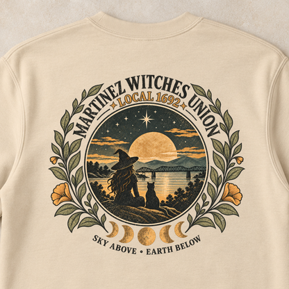 A cream-colored crewneck sweatshirt with a large graphic print on the back, laid flat on a light beige textured surface with small decorative pumpkins and dried flowers in the corners. The graphic features a circular design for the 'Nantucket Witches Union' with a witch silhouette, a full moon, and celestial elements.